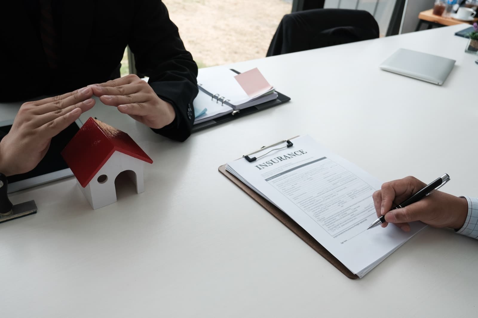 Hands protecting a model house beside an insurance document